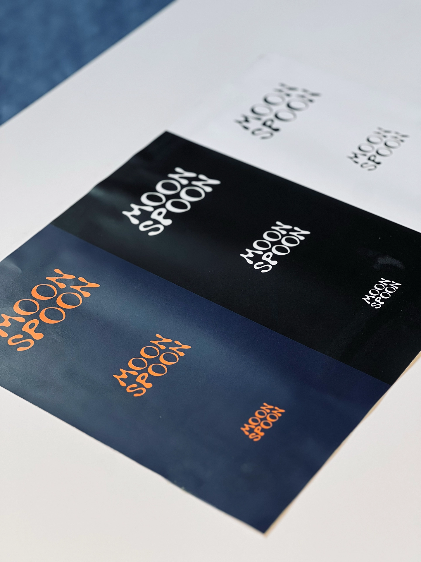 Logo-Farbvarianten des Schriftzugs „Moon Spoon" in verschiedenen Größen und Ausführungen: Schwarz auf Weiß, Weiß auf Schwarz und Orange auf Dunkelblau