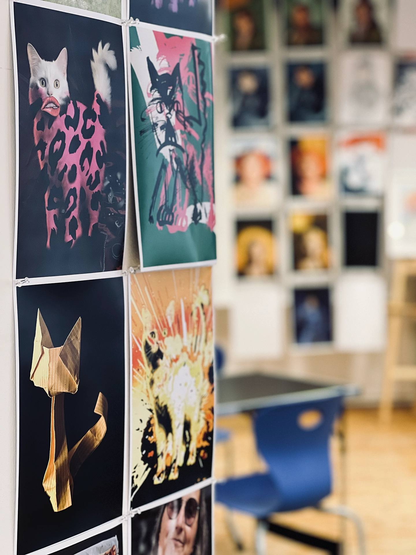Vier Katzen-Illustrationen in unterschiedlichen Techniken und Stilen an einer Wand: eine Fotomontage mit Leopardenmuster, eine expressive Zeichnung, eine Origami-Katze aus goldenem Papier und ein comicartiges Explosionsmotiv, im Hintergrund unscharf ein Raster aus Porträtdrucken