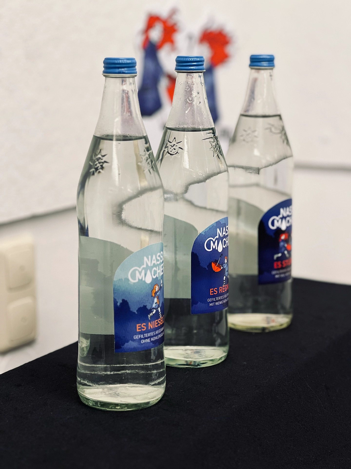 Drei Glasflaschen der Marke „Nassmacher" mit den Etiketten „Es nieselt", „Es regnet" und „Es stürmt", im Hintergrund unscharf Deko-Elemente der Kampagne