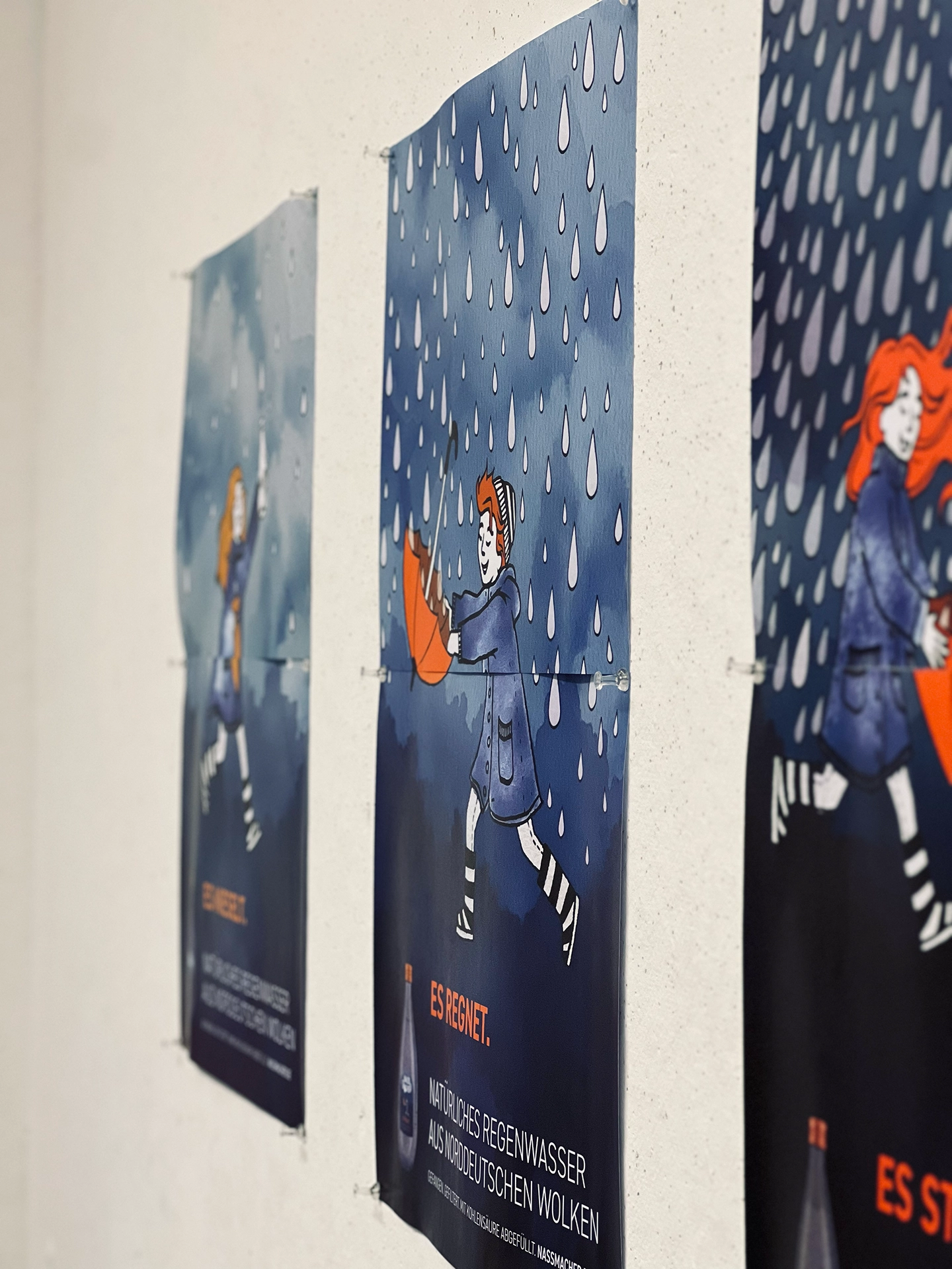 Reihe von Plakaten an einer Wand mit illustrierten Figuren im Regen und dem Slogan „Es regnet. Natürliches Regenwasser aus norddeutschen Wolken"