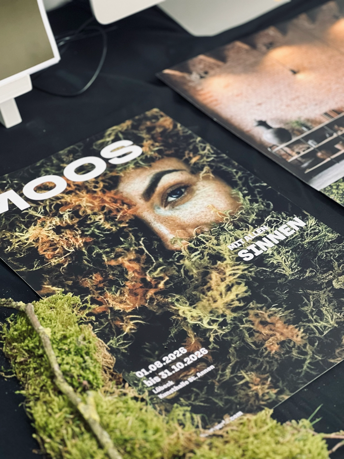 Ausstellungsplakat „Moos – mit allen Sinnen" mit einem in Moos eingebetteten Gesicht, daneben ein zweites Plakat, im Vordergrund echtes Moos