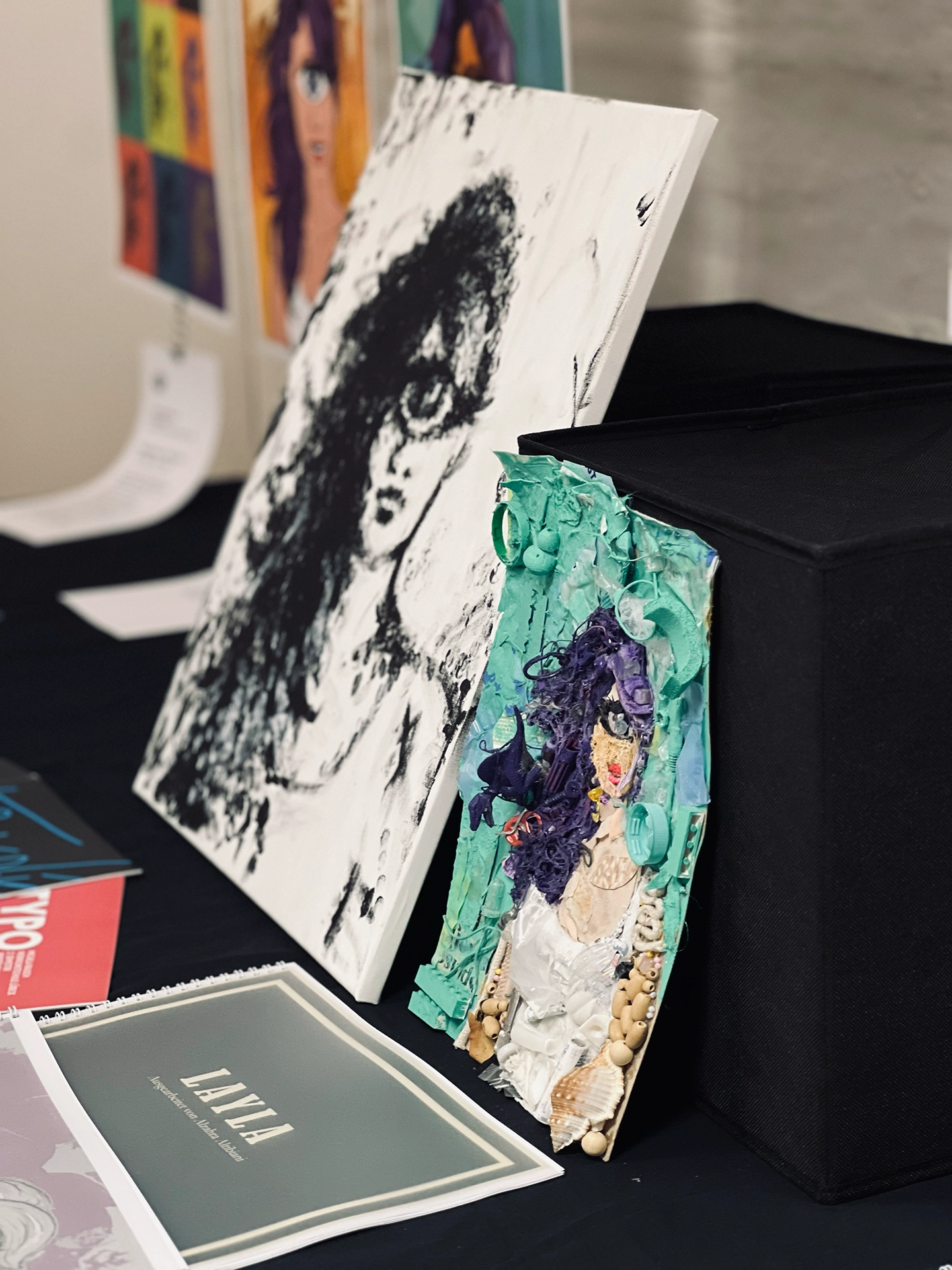 Ausstellungstisch mit einem monochromen gemalten Frauenporträt und einer türkisfarbenen Mixed-Media-Collage der Figur Leela aus Futurama, gestaltet aus Fundstücken.
