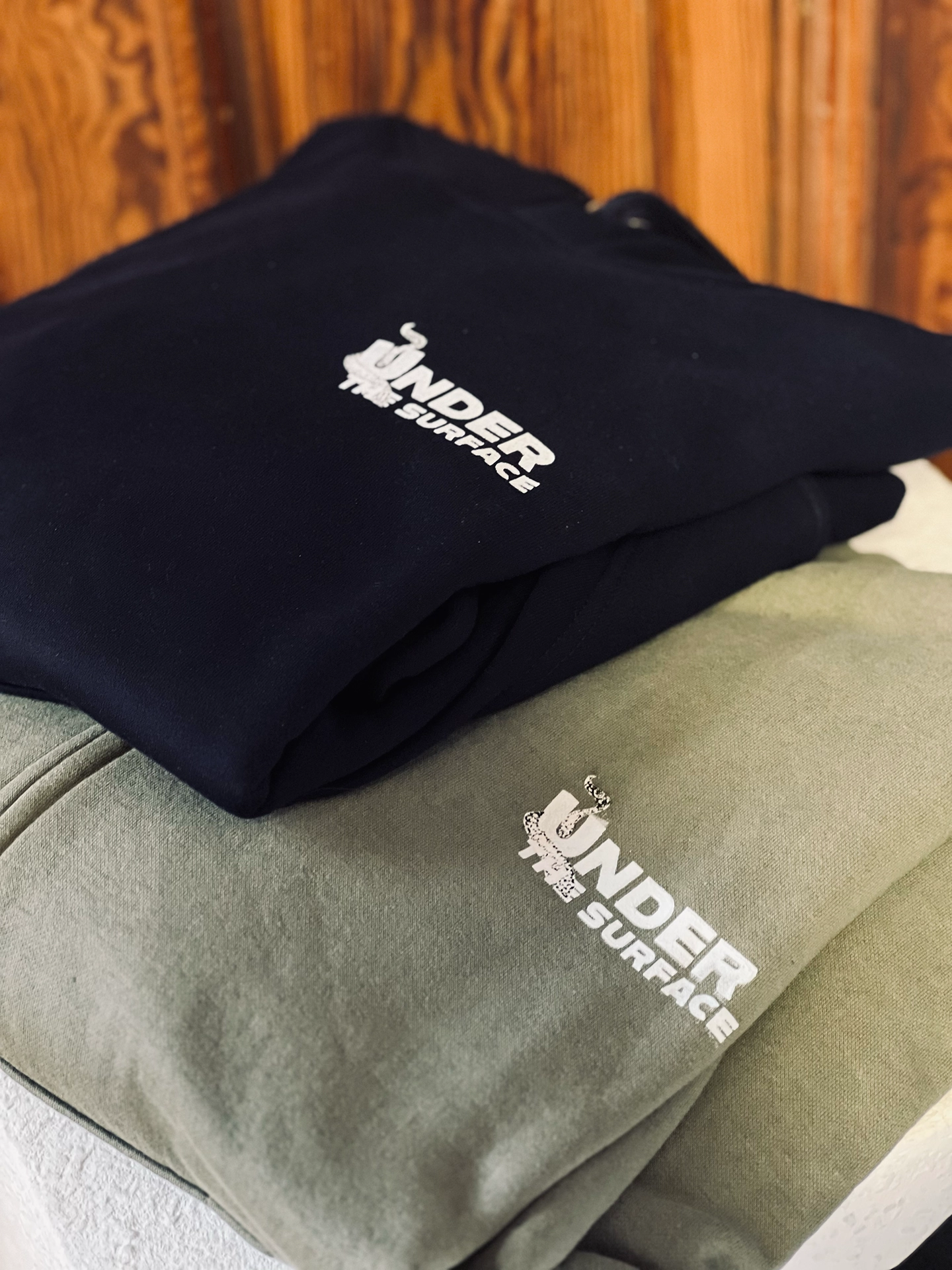 Zwei gefaltete Hoodies, dunkelblau und olivgrün, mit dem Aufdruck „Under the Surface"