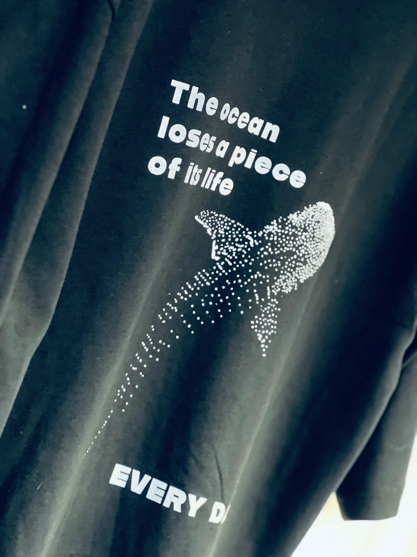 Nahaufnahme eines schwarzen T-Shirts mit dem Aufdruck „The ocean loses a piece of its life" und einem aus weißen Punkten gestalteten Walhai-Motiv
